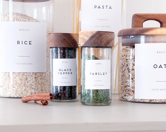 May include: A set of glass jars with wooden lids labeled with the words "Rice", "Black Pepper", "Parsley", and "Oats". The jars are filled with various pantry staples, including rice, black peppercorns, dried parsley, and oats. The labels are all white with black text and a thin black line underneath the text. The jars are arranged on a white countertop.