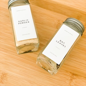May include: Two clear glass spice jars with silver lids on a wooden surface. One jar contains garlic powder, and the other contains bay leaves. Each jar has a white label with black text that reads "HERBS & SPICES", "GARLIC POWDER", "BAY LEAVES", and "100% NATURAL".