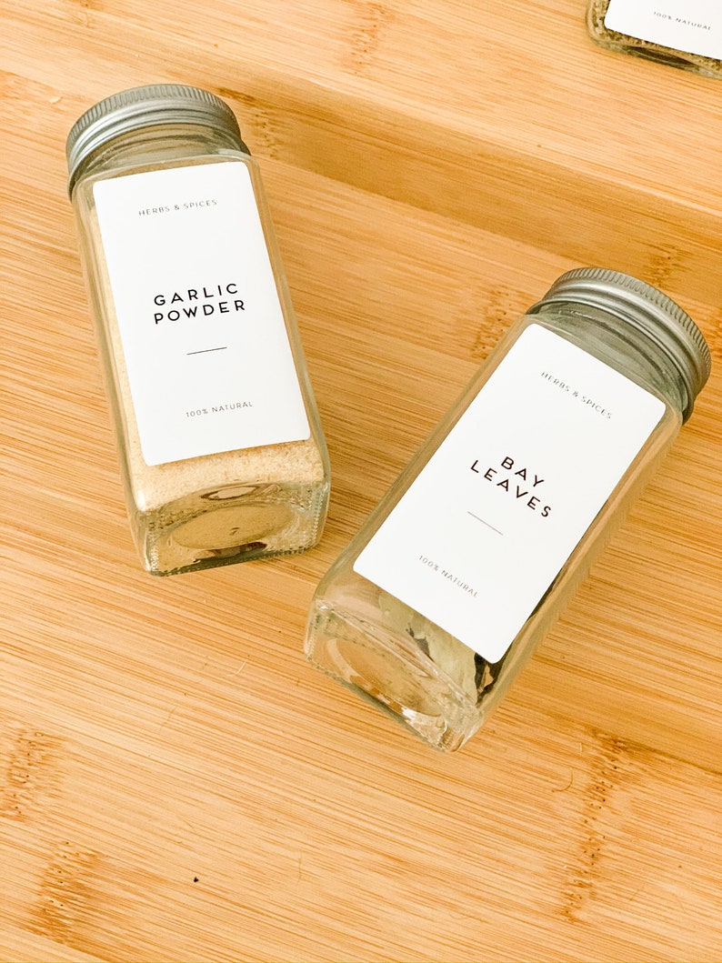 May include: Two clear glass spice jars with silver lids on a wooden surface. One jar contains garlic powder, and the other contains bay leaves. Each jar has a white label with black text that reads "HERBS & SPICES", "GARLIC POWDER", "BAY LEAVES", and "100% NATURAL".