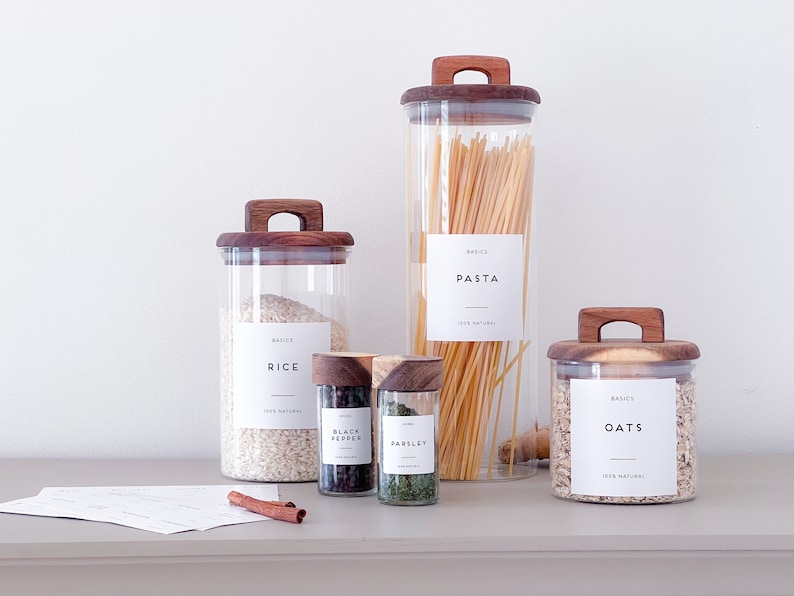 Set of glass jars with wooden lids in various sizes. On jars there are vinyl stickers that are printed with the name of the contents of the jars, flour, oats, rice, parsley.
