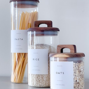 May include: Three glass jars with wooden lids and labels that say "BASICS" and the names of the contents: "PASTA", "RICE", and "OATS". Each jar contains a different type of dry good: pasta, rice, and oats.