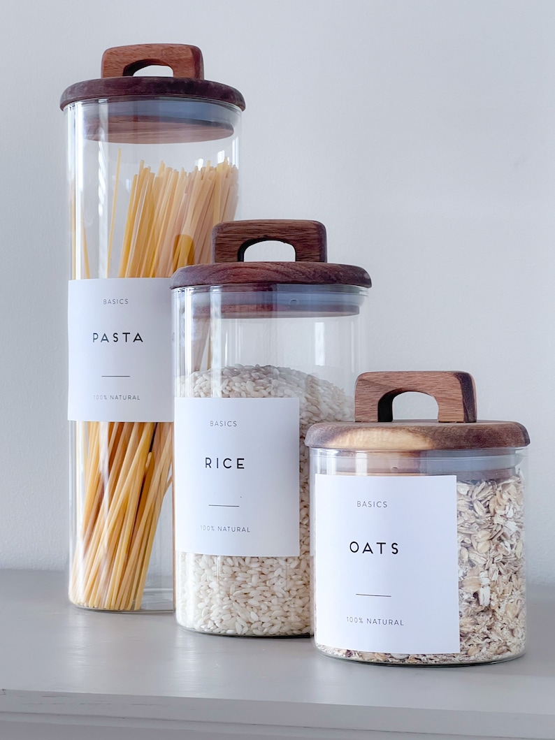 May include: Three glass jars with wooden lids and labels that say "BASICS" and the names of the contents: "PASTA", "RICE", and "OATS". Each jar contains a different type of dry good: pasta, rice, and oats.