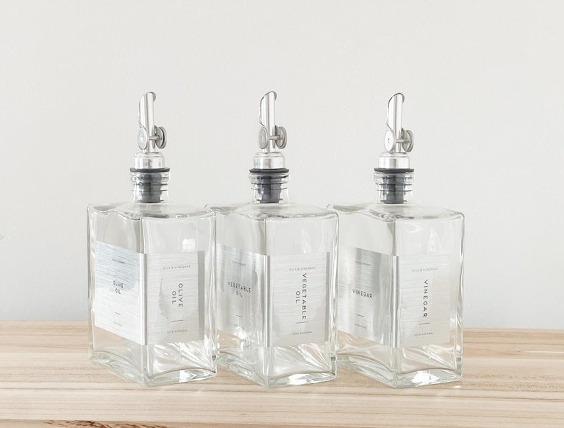May include: Three clear glass bottles with silver pour spouts and labels. The labels read "Olive Oil", "Vegetable Oil", and "Vinegar".