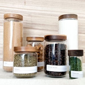 Könnte beinhalten: Eine Ansammlung von Gläsern mit Holzdeckeln, gefüllt mit verschiedenen Trockenprodukten. Die Gläser enthalten Zucker, Oregano, Kaffeebohnen und Petersilie. Jedes Glas hat ein weißes Etikett mit dem Inhalt.