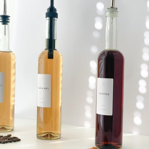 May include: Three glass bottles with pump tops filled with caramel, toffee, and vanilla syrups. The bottles are labeled with the flavor names. Two caramel candies are in the foreground.