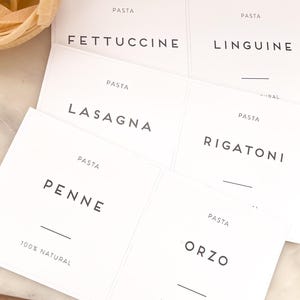 May include: White pasta labels with black text, including "Fettuccine," "Linguine," "Lasagna," "Rigatoni," "Penne," and "Orzo." The labels read "PASTA" at the top and "100% NATURAL" at the bottom. A bundle of uncooked fettuccine pasta is in the upper left.