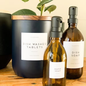 May include: A collection of cleaning supplies, including a dark blue canister labeled "Dish Washer Tablets" and amber glass bottles for "Dish Soap" and "Hand Soap." Each container has a black pump and a white label with black text. The products are displayed on a wooden surface.