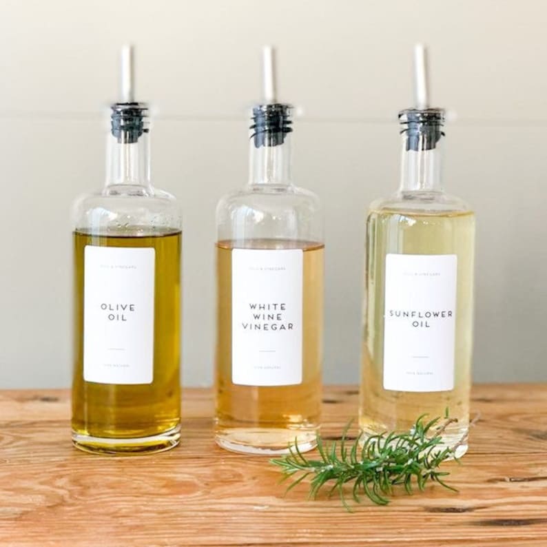 May include: Three glass bottles with black lids and labels. The bottles contain olive oil, white wine vinegar, and sunflower oil. The labels are white with black text. The bottles are sitting on a wooden surface with a sprig of rosemary in front of them.