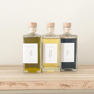 May include: Three glass bottles with cork tops and labels. The bottles contain olive oil, white vinegar, and soy sauce. The labels are white with black text.