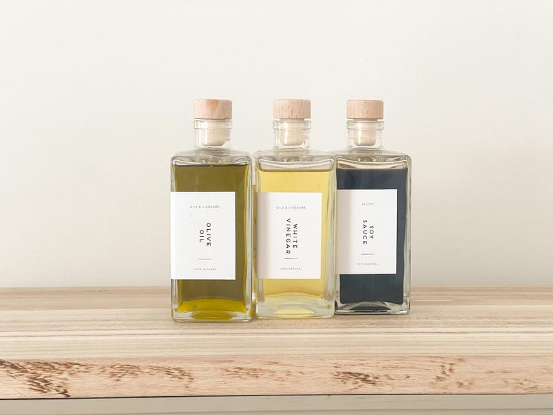 May include: Three glass bottles with cork tops and labels. The bottles contain olive oil, white vinegar, and soy sauce. The labels are white with black text.