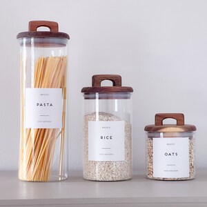 May include: Three glass jars with wooden lids and labels. The jars contain pasta, rice, and oats. The labels are white with black text and say "BASICS" and the name of the food.