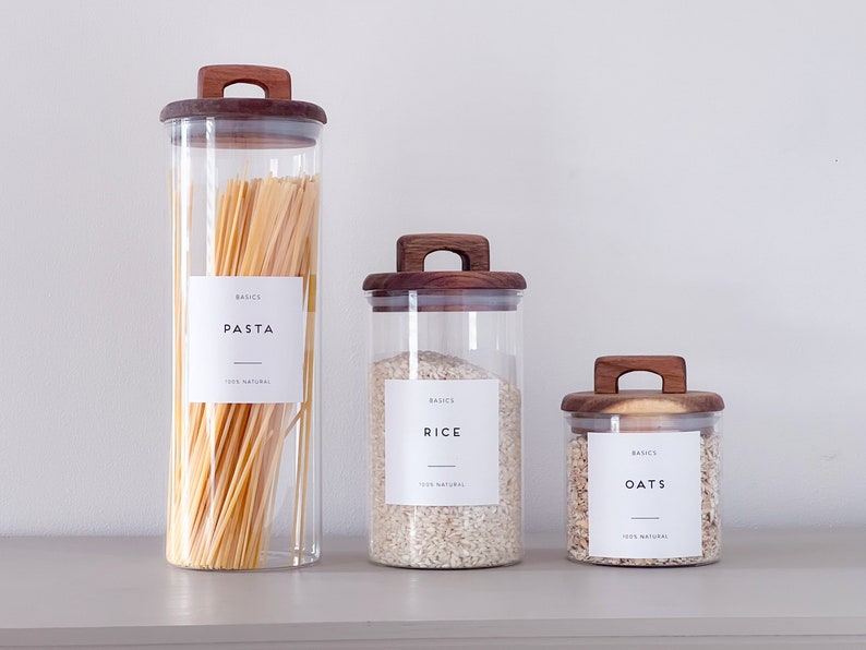 May include: Three glass jars with wooden lids and labels. The jars contain pasta, rice, and oats. The labels are white with black text and say "BASICS" and the name of the food.