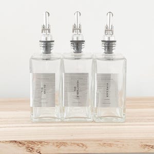 May include: Three clear glass bottles with silver tops and labels. The labels read "Olive Oil", "Vegetable Oil", and "Vinegar".