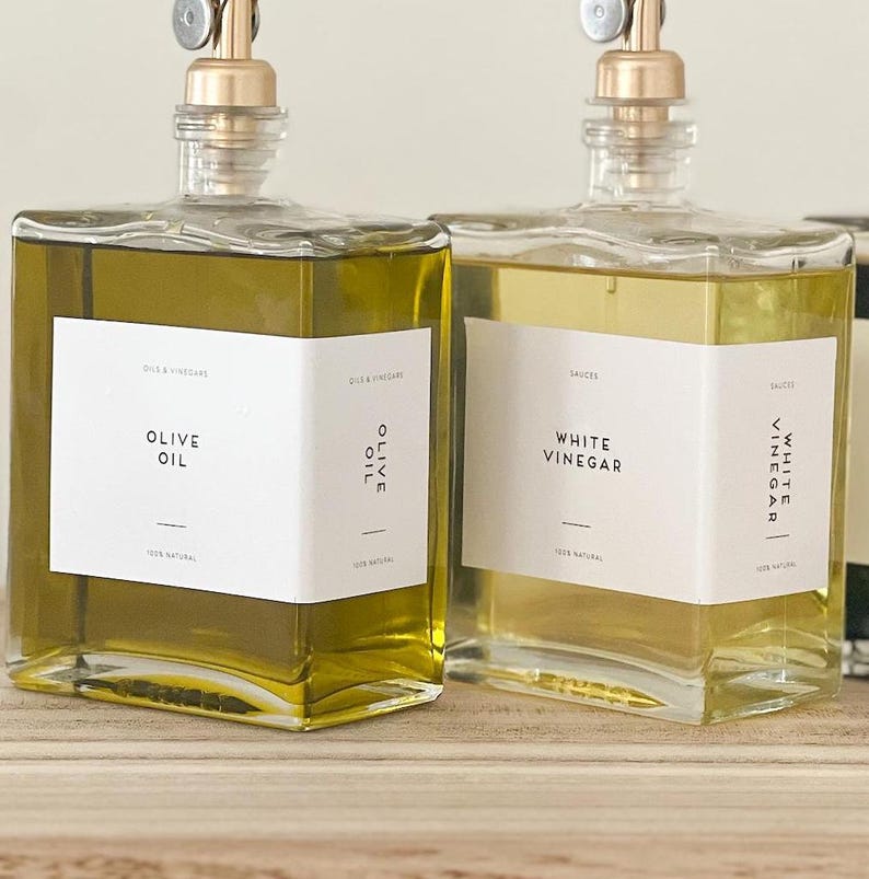 May include: Two clear glass bottles with gold tops and labels. The bottle on the left is filled with olive oil and the label reads "Olive Oil". The bottle on the right is filled with white vinegar and the label reads "White Vinegar".