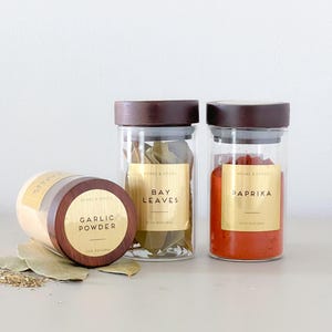 May include: Three glass spice jars with wooden lids. One jar is labelled "Garlic Powder" and is lying on its side. The other two jars are upright and labelled "Bay Leaves" and "Paprika". The jars are on a white surface.