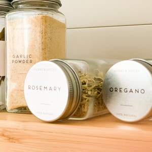 May include: A selection of clear glass spice jars with metal lids, filled with herbs and spices, including bay leaves, garlic powder, rosemary, and oregano. The jars are labelled with "Herbs & Spices" and "100% Natural" on a wooden shelf.