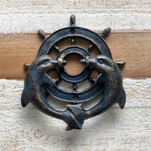 May include: A maritime door knocker with a ship's wheel design and two dolphins. The dolphins are on either side of the wheel, as if holding it. The knocker has a distressed blue and gold finish, giving it a vintage appearance.