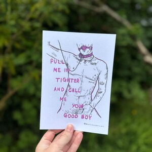 May include: A black and white illustration of a person with a pink gag in their mouth and a pink collar around their neck. The person is wearing a pink choker and has a pink ring on their finger. The text "PULL ME IN TIGHTER AND CALL ME YOUR GOOD BOY" is written in pink letters on the white background.