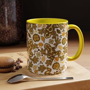 May include: White ceramic mug with a yellow interior and handle. The mug has a repeating pattern of brown frogs, leaves, and flowers. A spoon and a pastry are on a wooden surface next to the mug.