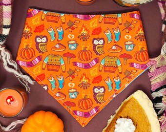 Op de afbeelding: Een driehoekige bandana voor huisdieren met een oranje achtergrond en een herfstpatroon van pompoenen, uilen, truien, kampvuren, mokken en sokken. De stof wordt omringd door een geruite deken, twee oranje kaarsen en een stuk pompoentaart.