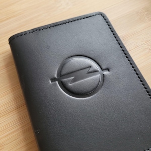 May include: Black leather wallet with a debossed Opel logo. The wallet has a rectangular shape with rounded corners and a stitched border. The Opel logo is a circle with a lightning bolt through it.