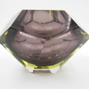 K&ouml;nnte beinhalten: Ein facettierter Aschenbecher aus Glas mit geometrischer Form. Der Aschenbecher besteht aus klarem Glas mit einer violetten T&ouml;nung und einem gelben Rand.