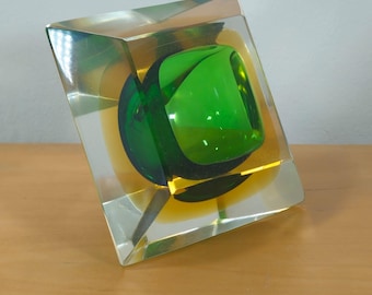 May include: A square glass sculpture with a green sphere inside. The sphere is surrounded by a clear glass layer and a yellow glass layer.