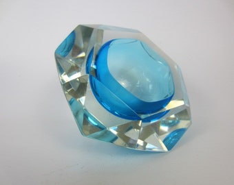 May include: A clear glass ashtray with a faceted design and a bright blue center. The ashtray is shaped like a hexagon.