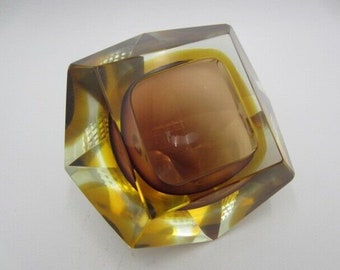 May include: A geometric glass ashtray with a clear outer layer and a brown inner layer. The ashtray has a square shape with beveled edges.