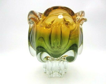 May include: A green and amber glass vase with a unique, organic shape. The vase has a wide, open mouth and a narrow base. It is made of clear glass with a green and amber color gradient.