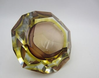 May include: A faceted, octagonal ashtray in shades of amber, brown, and yellow. The ashtray has a central bowl and a geometric design, reflecting light. The glass ashtray is a decorative item.