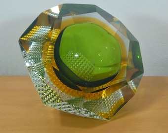 May include: A faceted glass decorative object with a central green orb and yellow and clear accents. The geometric design features a textured pattern on the sides and a polished finish, creating a striking display piece. The object is approximately 6 inches wide.