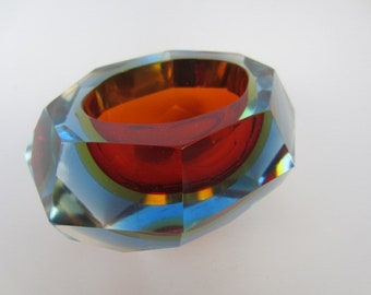 May include: A multi-colored glass ashtray with a geometric design. The ashtray is made of clear glass with blue, green, and orange accents. The inside of the ashtray is red.