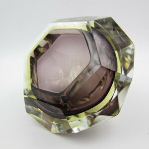 K&ouml;nnte beinhalten: Ein facettierter Aschenbecher aus Glas mit geometrischer Form. Der Aschenbecher ist aus klarem Glas mit einer violetten T&ouml;nung und hat einen gelben Rand.