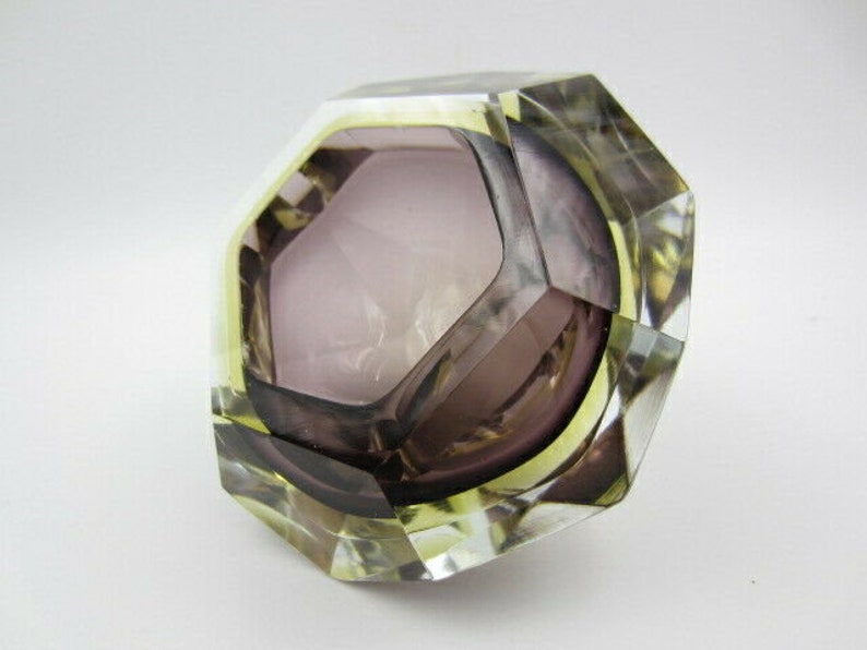 K&ouml;nnte beinhalten: Ein facettierter Aschenbecher aus Glas mit geometrischer Form. Der Aschenbecher ist aus klarem Glas mit einer violetten T&ouml;nung und hat einen gelben Rand.
