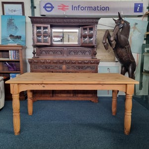 May include: A light-coloured wooden dining table with four turned legs. The rectangular tabletop is made of planks. The table is set on a blue carpet. In the background is a dark wooden cabinet and a large wooden horse statue.