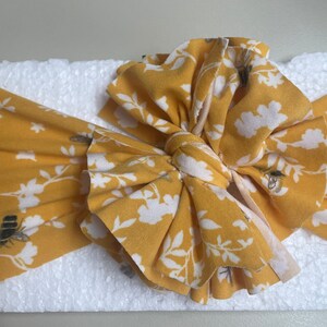 May include: A yellow headband with a white floral pattern and small bee designs. The headband is tied into a large bow.