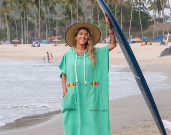 Op de afbeelding: Een vrouw in een groene badjas met capuchon met kleurrijke strepen en een brede strohoed staat op een zandstrand en houdt een blauw surfboard vast.