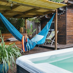 Op de afbeelding: Een turquoise hangmat hangt onder een houten pergola, met een houten hut op de achtergrond. Een jacuzzi staat op de voorgrond. Een deken en kussen liggen in de hangmat. Planten en lichtslingers dragen bij aan de buitenscène.