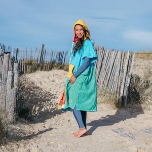 May include: A person wearing a hooded surf poncho in shades of turquoise and green, with a yellow hood and a colorful pocket. The poncho is worn over a long-sleeved blue shirt and blue leggings. The background features a wooden fence and sand.