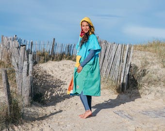 Könnte beinhalten: Eine Person trägt einen Kapuzen-Surfponcho in Türkis und Grün mit einer gelben Kapuze und einer bunten Tasche. Der Poncho wird über einem langärmeligen blauen Shirt und blauen Leggings getragen. Im Hintergrund ein Holzzaun und Sand.