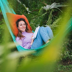 Może przedstawiać: Osoba relaksuje się w turkusowym i zielonym hamaku z pomarańczową poduszką. Osoba ma na sobie lawendowy golf i niebieskie dżinsy, trzymając książkę. Bujne zielone liście otaczają hamak, tworząc spokojną scenerię na świeżym powietrzu.