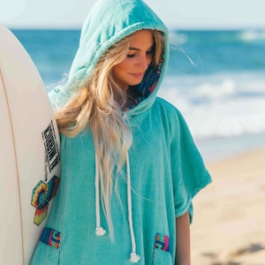 Peut inclure: Une femme portant une serviette à capuche turquoise avec des bordures colorées se tient sur une plage de sable en tenant une planche de surf blanche avec un motif coloré.