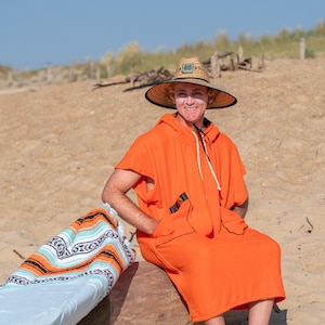 May include: A bright orange hooded poncho with a front pocket and white drawstring, paired with a wide-brimmed straw hat featuring a square patch. Beside the poncho lies a folded blanket with a white, light blue, orange, and black pattern.