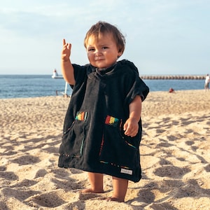 May include: A child wearing a black hooded beach poncho with rainbow accents on the pockets stands on a sandy beach. The poncho has a small tag with text. The child is barefoot and raising their hand. The background includes the ocean and a clear sky.