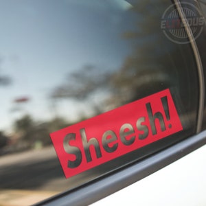 May include: A red rectangular sticker with the word "Sheesh!" cut out. The sticker is affixed to a car window, with the background showing a blurred outdoor scene.