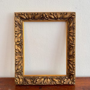 May include: A rectangular, ornate gold picture frame. The frame features a detailed leaf and scroll design around the edges. The frame is empty, with a visible opening in the center. The frame is sitting on a wooden surface.
