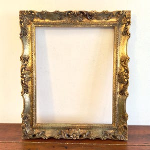 Puede incluir: Marco de fotos dorado y ornamentado con tallas detalladas. El marco tiene una abertura rectangular y está colocado contra una pared blanca y una superficie de madera. El diseño del marco presenta intrincados patrones florales y de volutas.