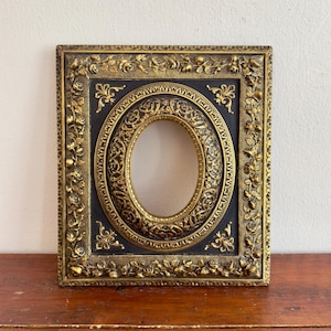 Puede incluir: Un marco de fotos rectangular con un centro negro y una abertura ovalada. El marco presenta detalles en relieve de color dorado, incluyendo motivos de rosas a lo largo del borde exterior y volutas intrincadas alrededor de la abertura ovalada.
