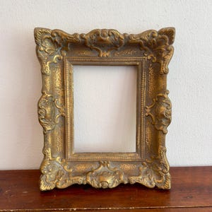 Può includere: Una cornice rettangolare con finitura dorata anticata. La cornice presenta volute ornamentate in rilievo e motivi a forma di conchiglia lungo i bordi, che circondano un'apertura centrale rettangolare con una superficie testurizzata e usurata.